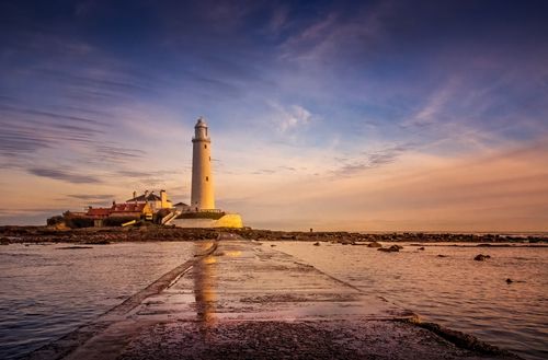 Free A picturesque lighthouse stands tall by the sea during a tranquil sunset, reflecting on the water. Stock Photo