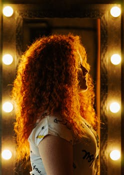 A side view portrait of a redhead woman with curly hair posing by a vanity mirror with lights.