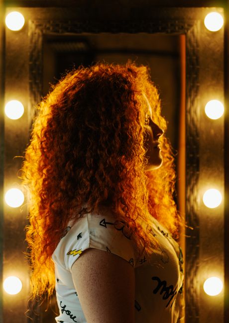 A side view portrait of a redhead woman with curly hair posing by a vanity mirror with lights.