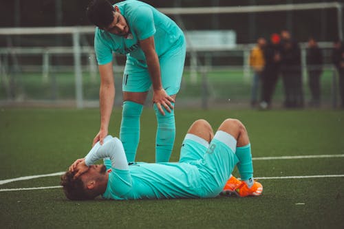 Free A soccer player lying on the grass field being consoled by a teammate in sportswear. Stock Photo