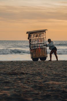 A vendor pushes a cart of jewelry along the beach at sunset with the ocean in the background.