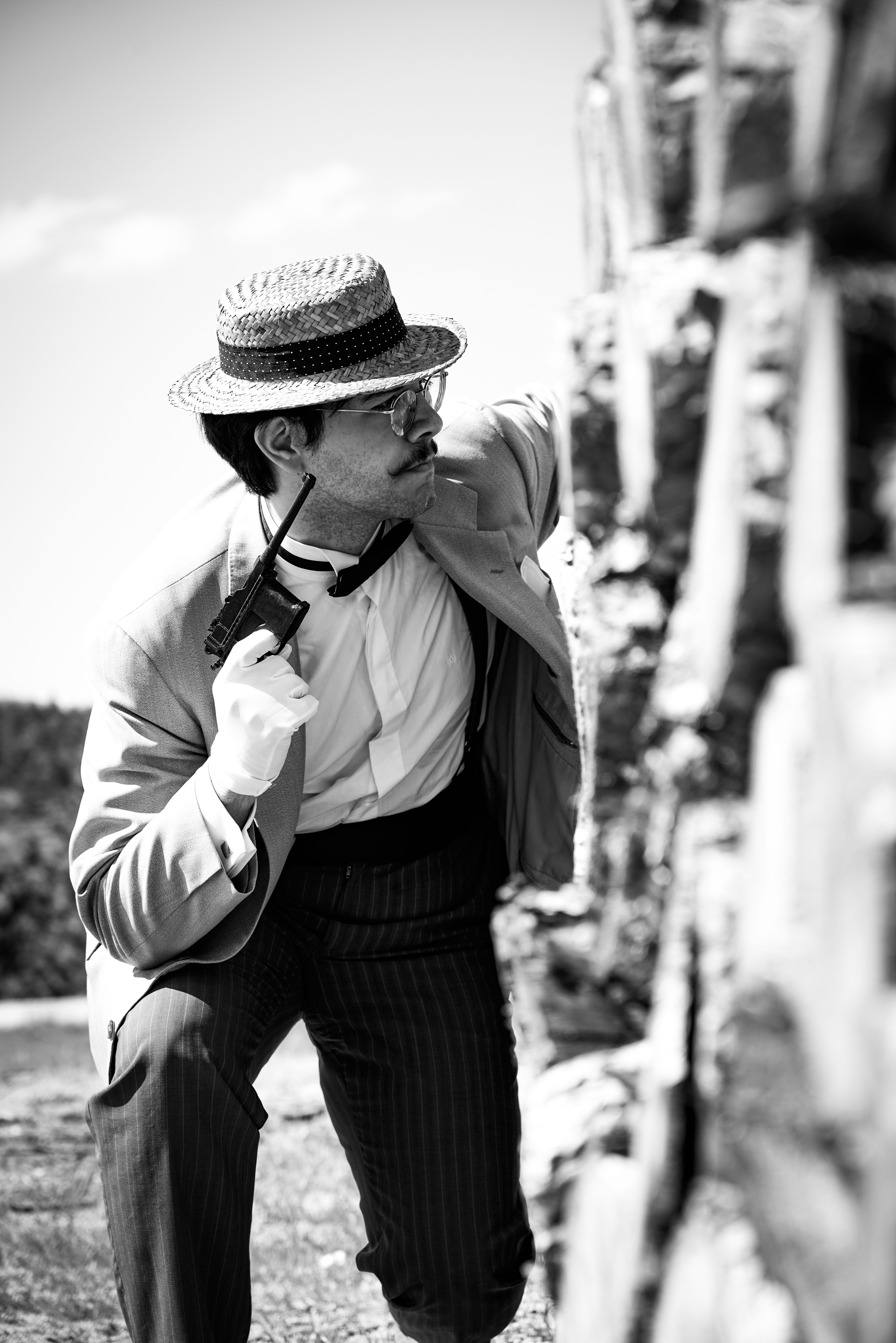 Black and white photo of a man in vintage attire and eyeglasses holding a pistol against a wall outdoors.