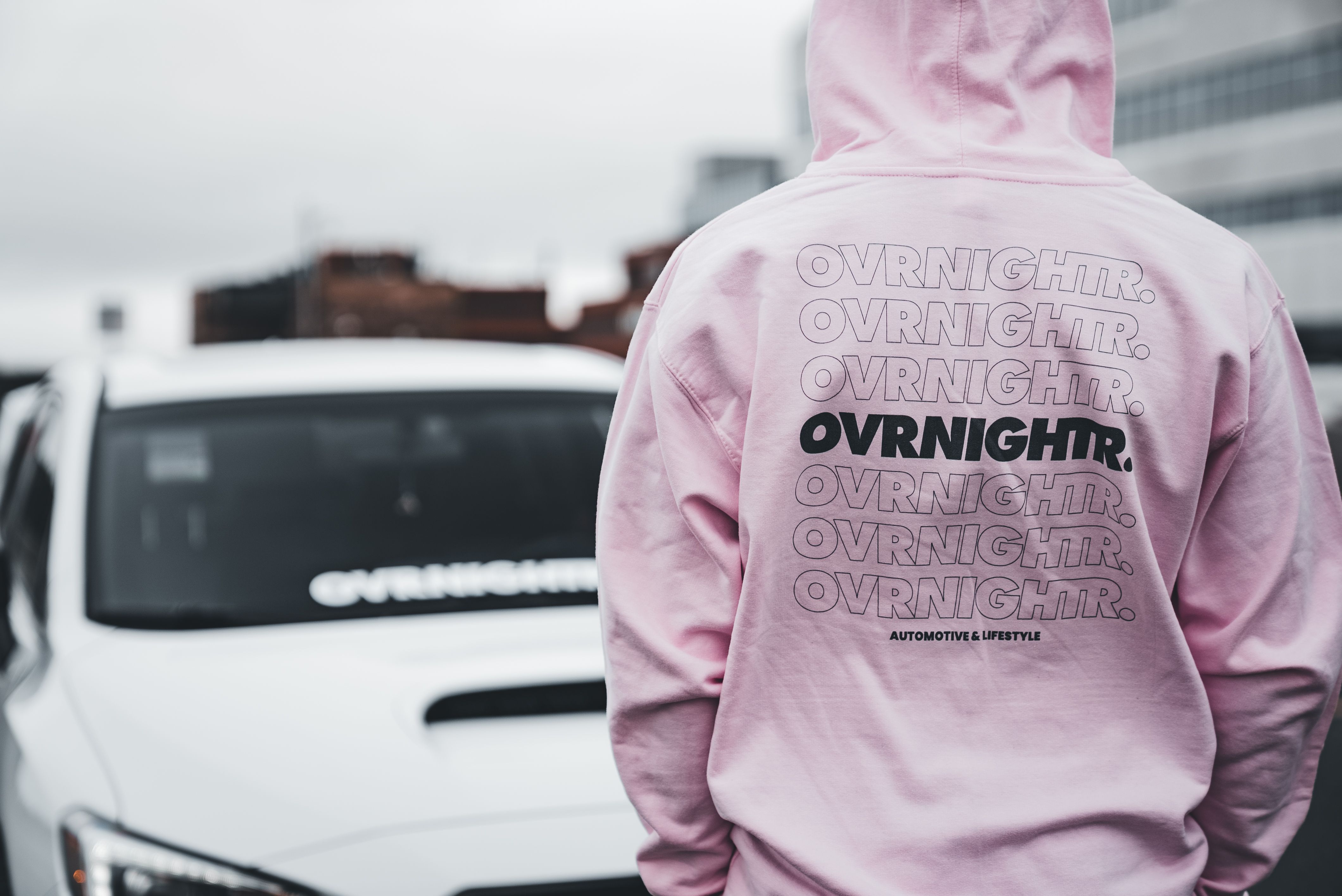 Stylish back view of person in pink hoodie standing near white sports car in urban setting.