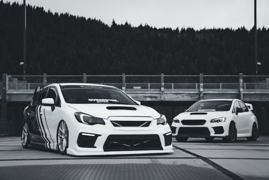Two modified white sports cars parked outdoors near a forest in an urban setting.