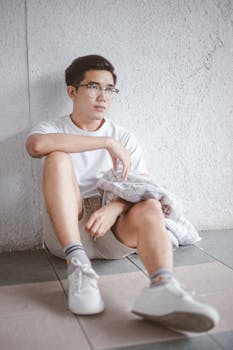 Asian man with eyeglasses sitting casually against a white wall in casual fashion.
