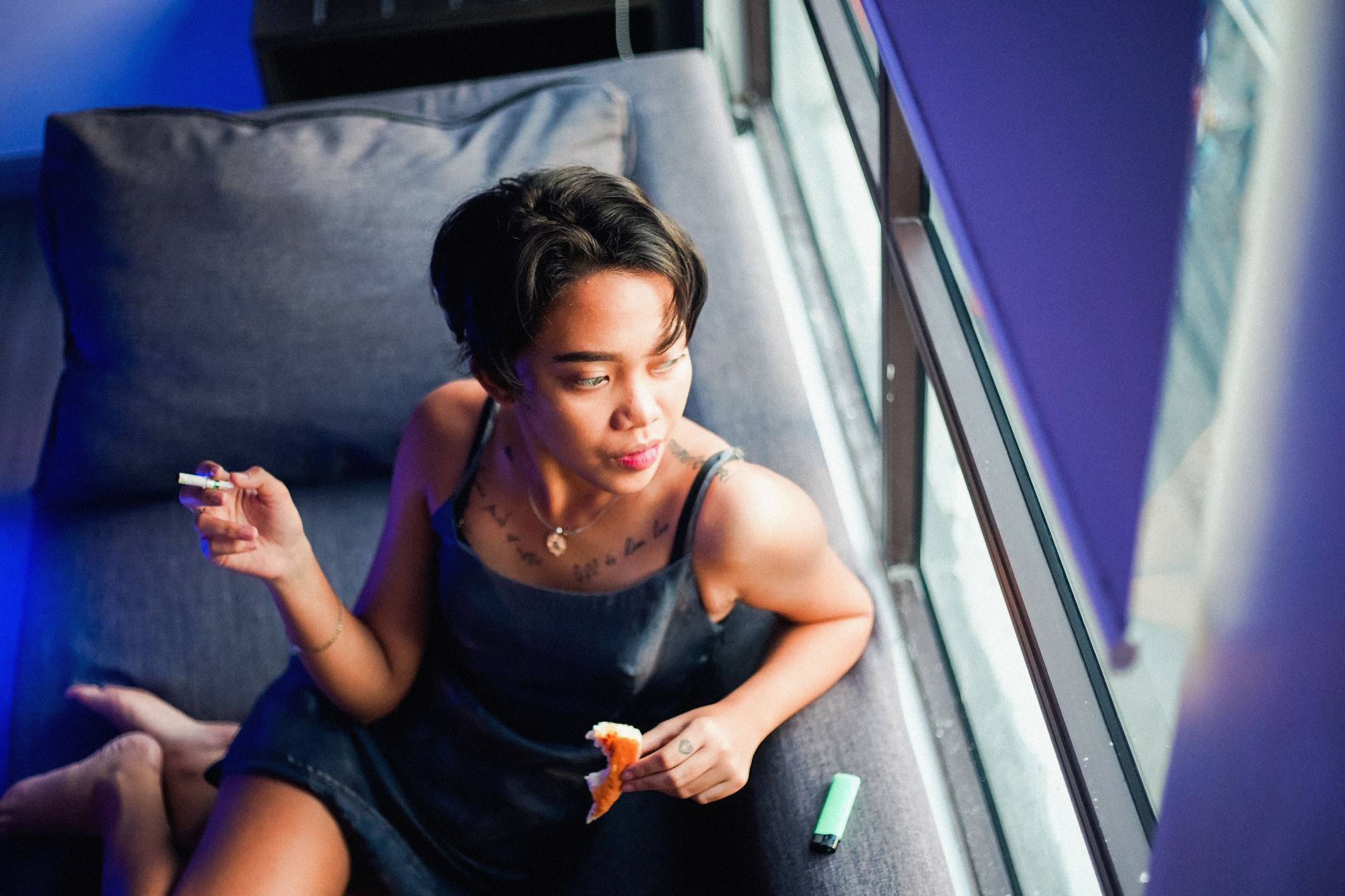 A young woman with short hair sits by the window, enjoying a cigarette indoors.