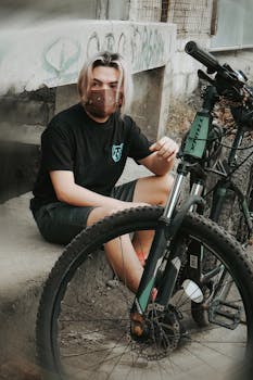 Young man with dyed hair sits with a bicycle, wearing a face mask in an urban setting.
