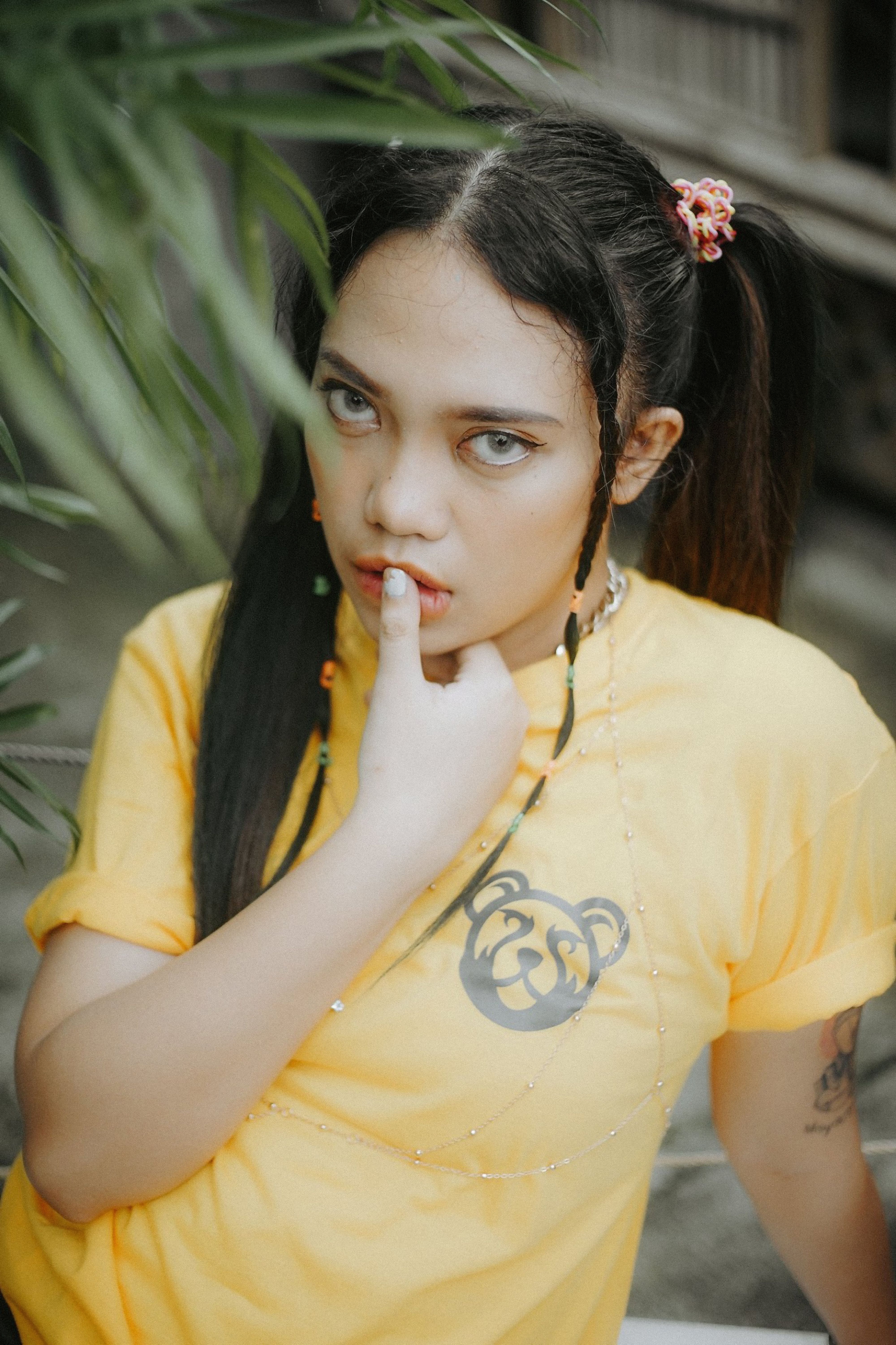 A young woman with tattoos and a yellow t-shirt posing outdoors among plants.