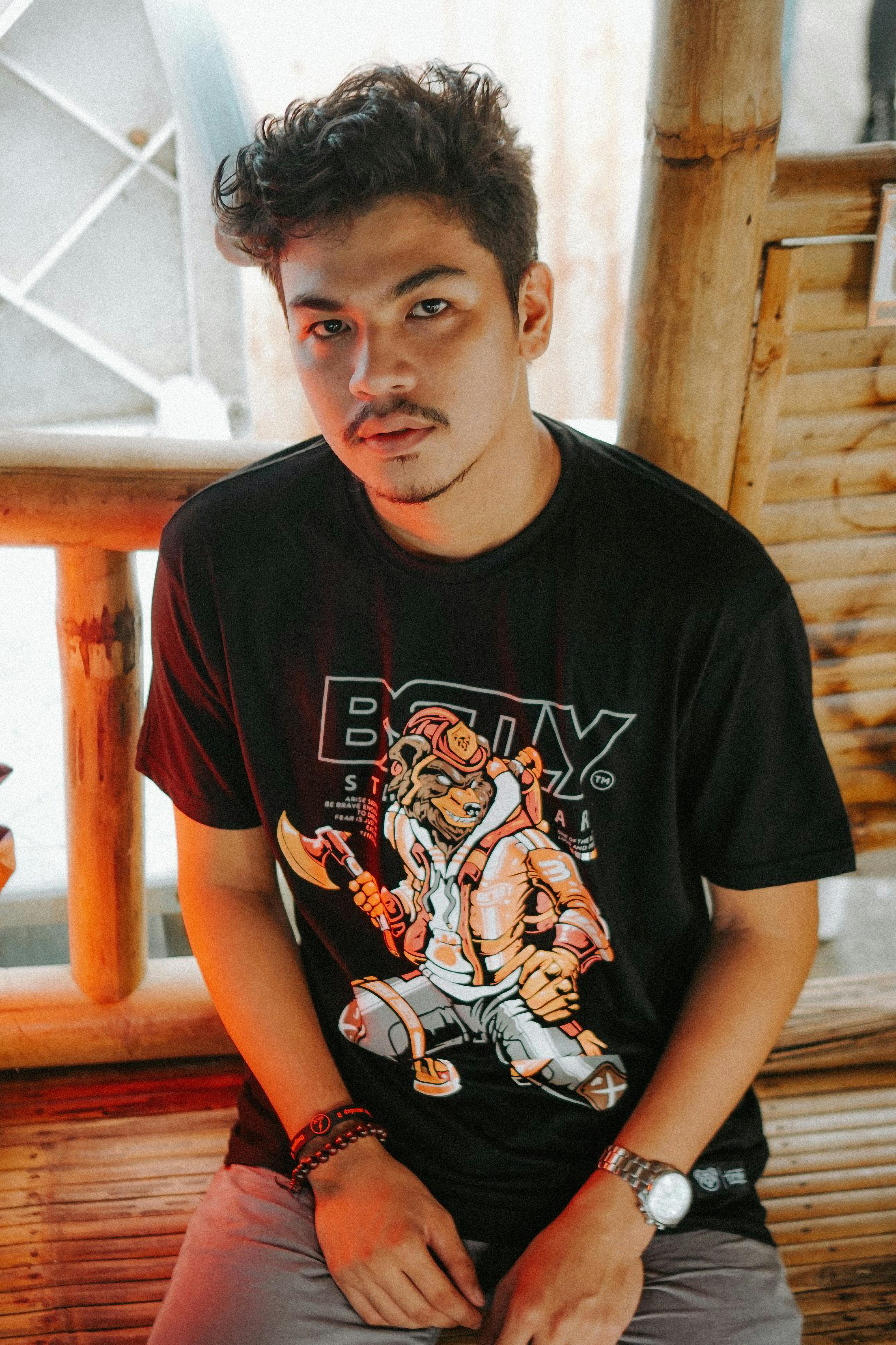 Young man with styled hair wearing a graphic t-shirt, sitting indoors on a bamboo bench.