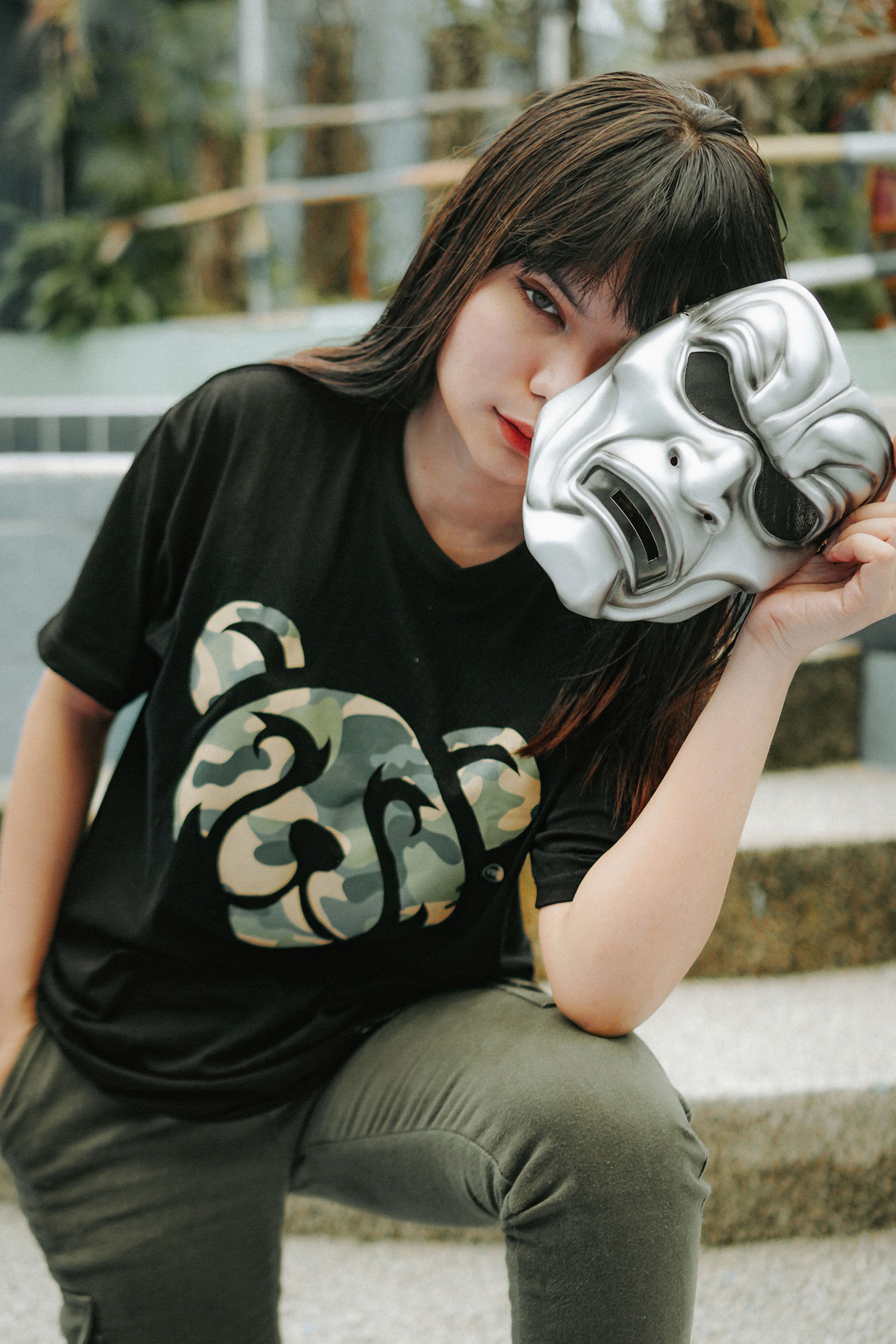 Stylish woman in graphic T-shirt posing with a dramatic silver mask on outdoor steps.