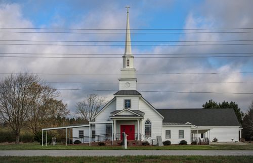 Free A classic white church with a red door and tall steeple under a blue sky, symbolizing spirituality and tradition. Stock Photo