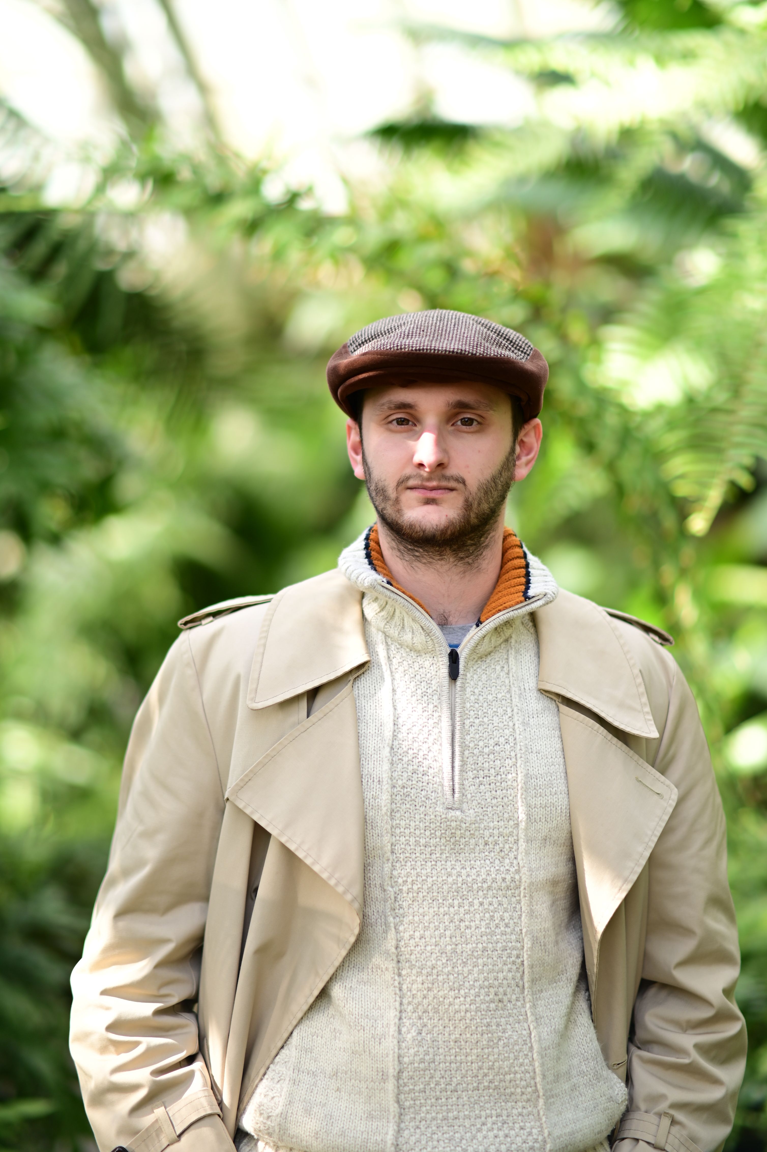 Portrait of a man in a trench coat and cap surrounded by lush greenery.