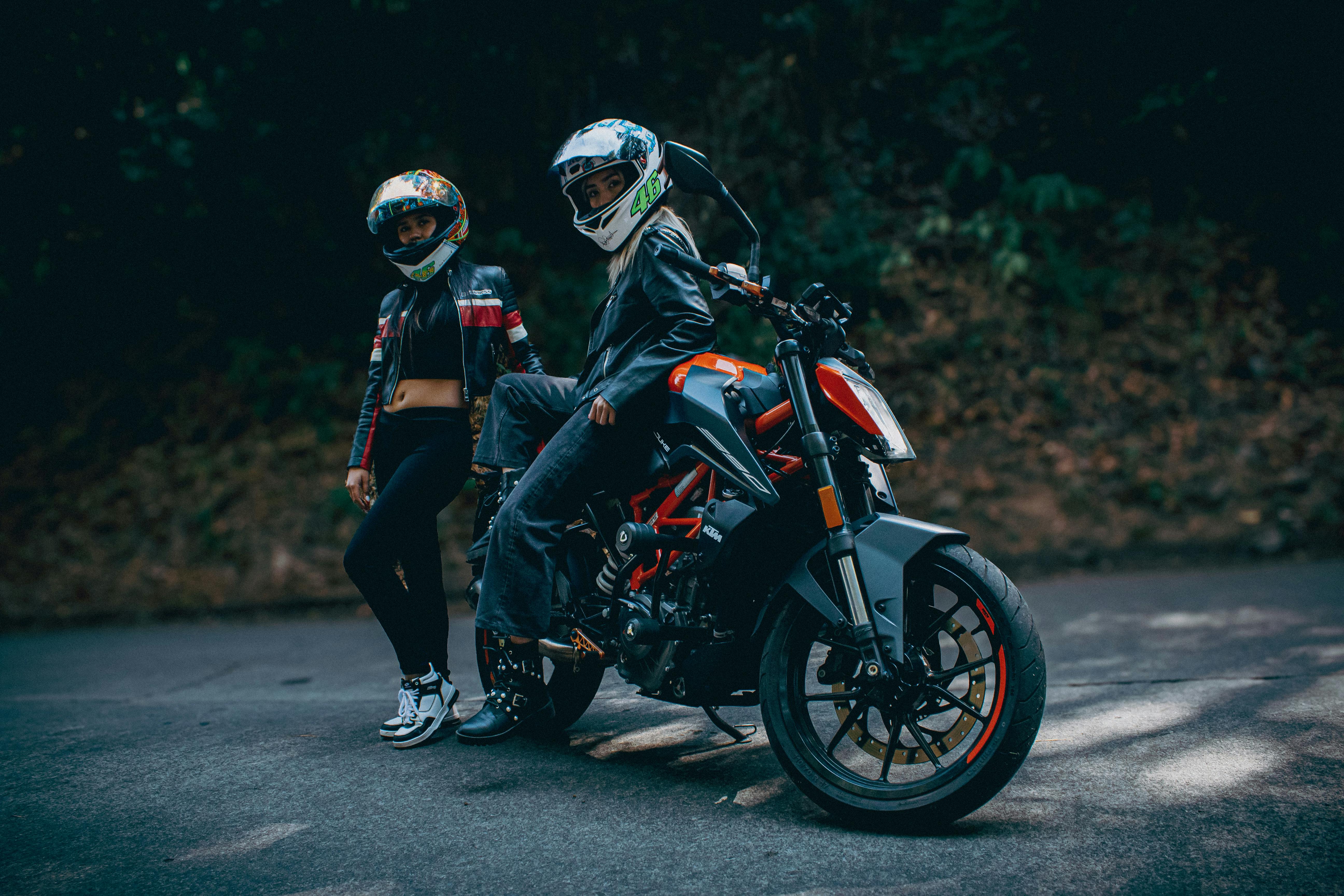 Two bikers wearing helmets and leather jackets posed with a KTM motorcycle on a rural road.