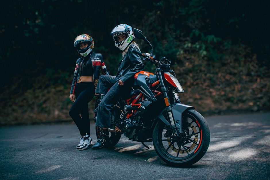 Two bikers wearing helmets and leather jackets posed with a KTM motorcycle on a rural road.