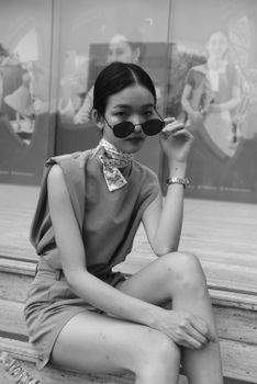 Black and white portrait of a fashionable woman sitting outdoors with sunglasses and a scarf.