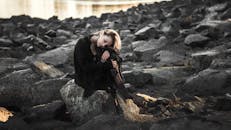 一名女子身穿黑衣，若有所思地坐在水边的岩石上