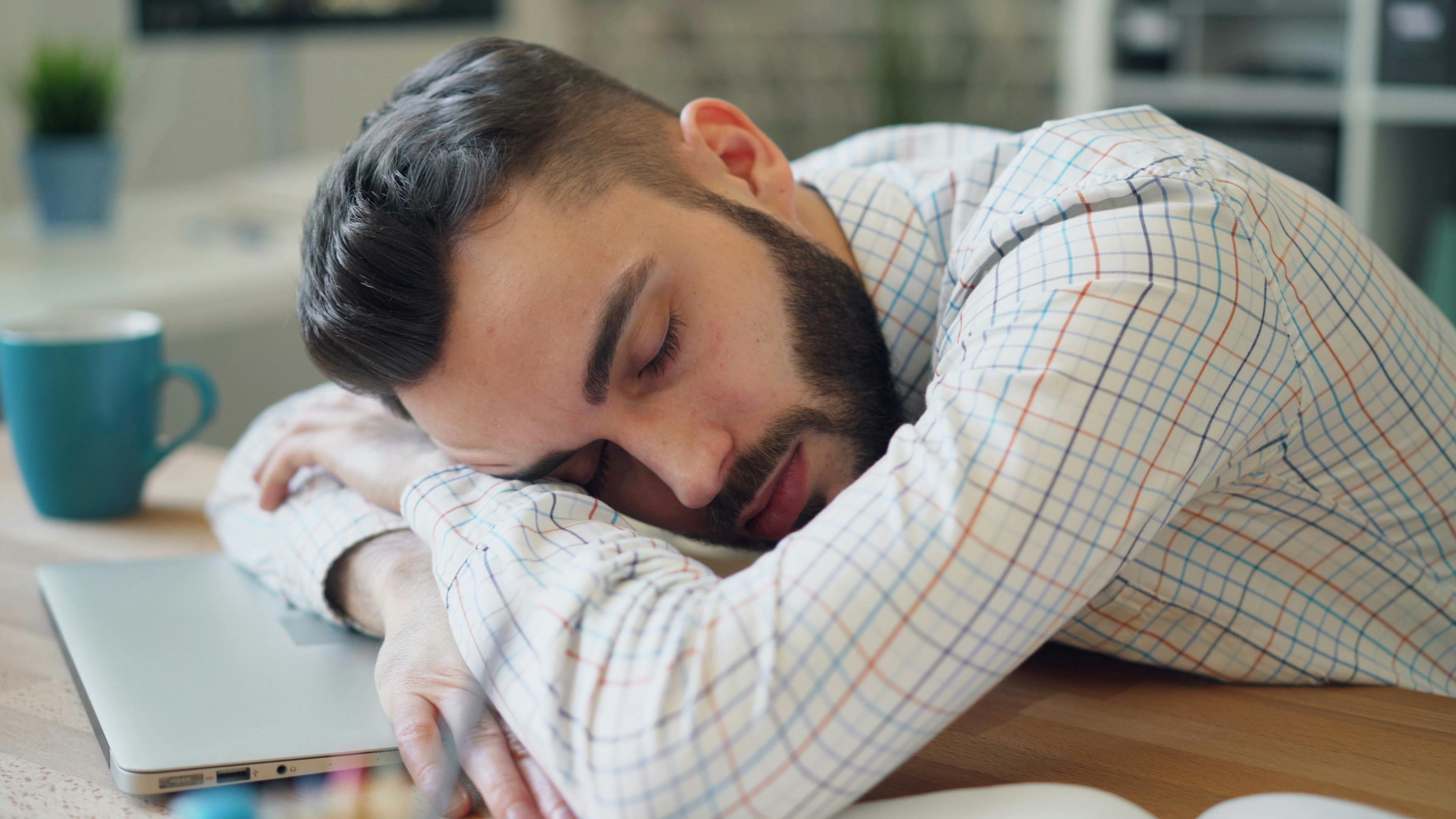 Man with a beard resting on a desk next to a laptop, appearing exhausted in an office setting.