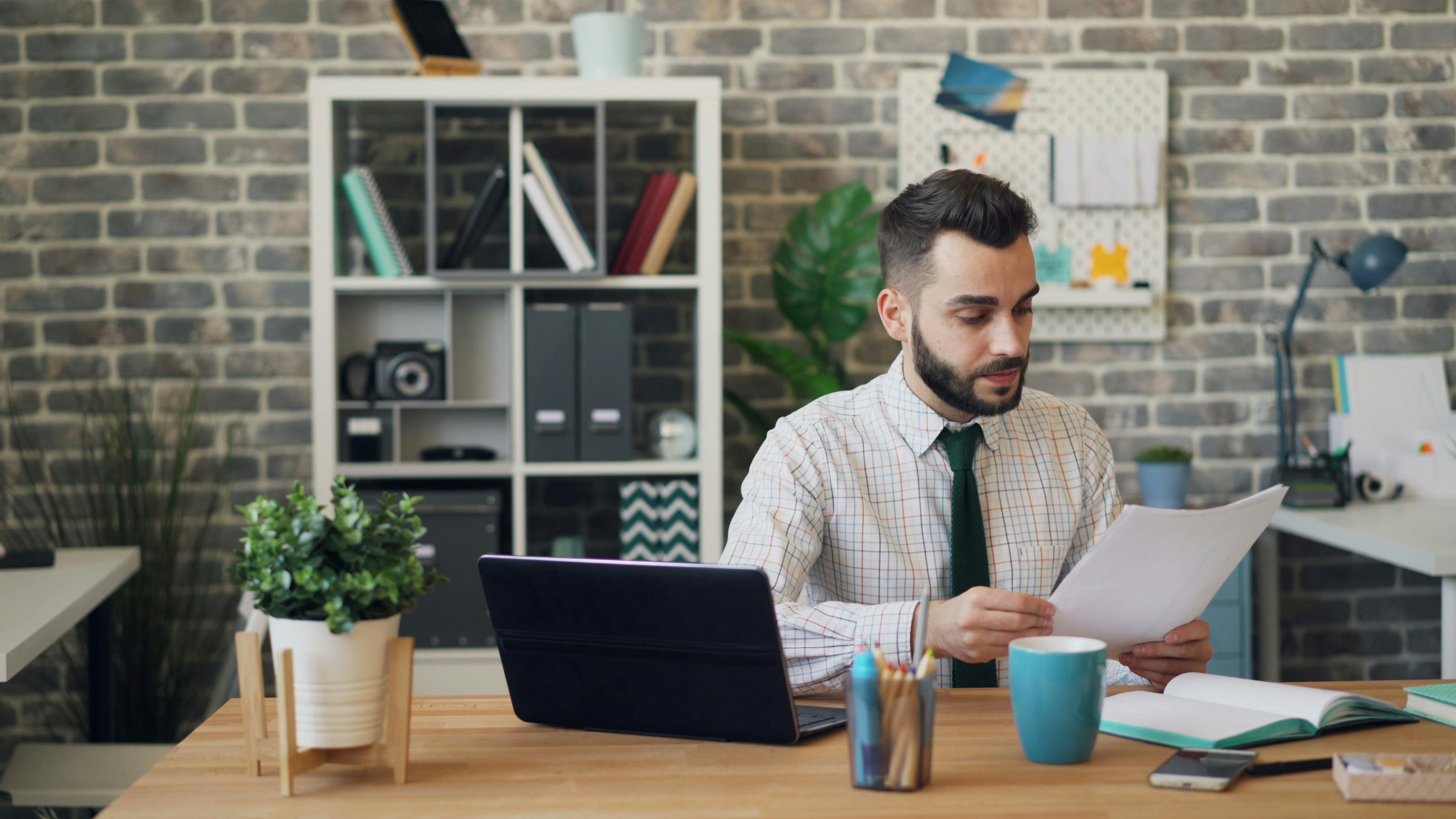 Young man reading papers at desk with laptop and office decor, focused on work.