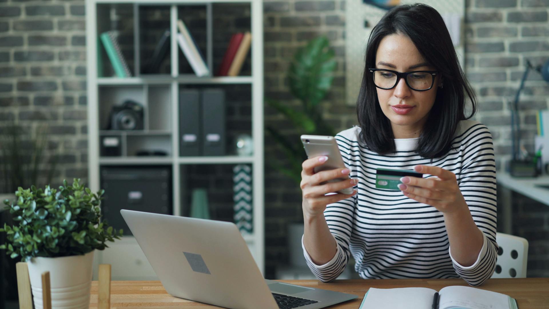 Professional woman using phone and credit card for online transaction at desk in office.