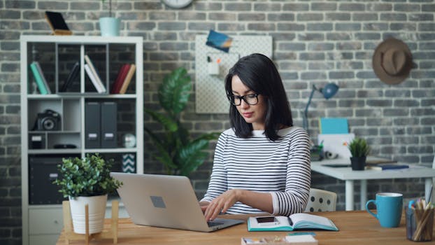 Woman with glasses working on a laptop in a stylish modern office with a brick wall background.