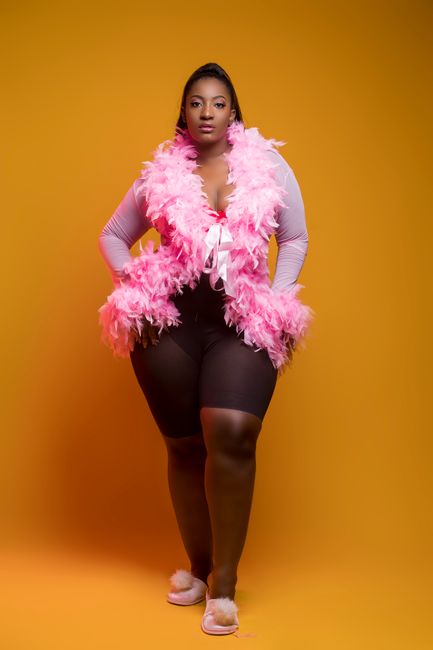 African woman in pink feathered outfit posing confidently on vibrant orange backdrop.