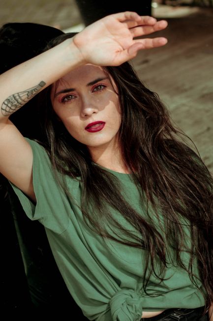 A fashionable young woman poses outdoors with a confident expression, wearing a green top.