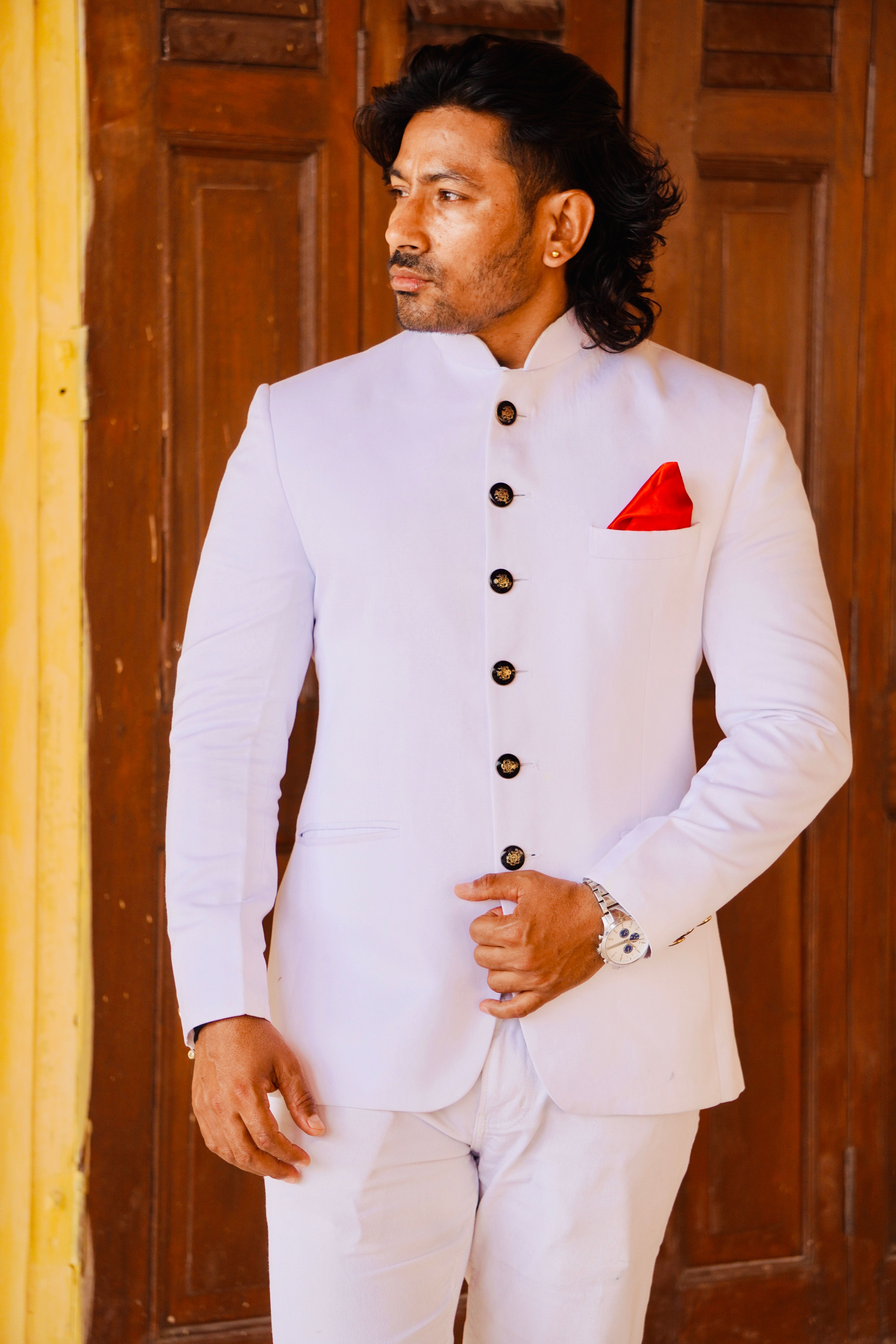 A man in a stylish white suit and red pocket square standing by a wooden door.