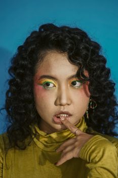 Portrait of a young woman with curly hair, colorful makeup, and a yellow top against a vibrant blue background.