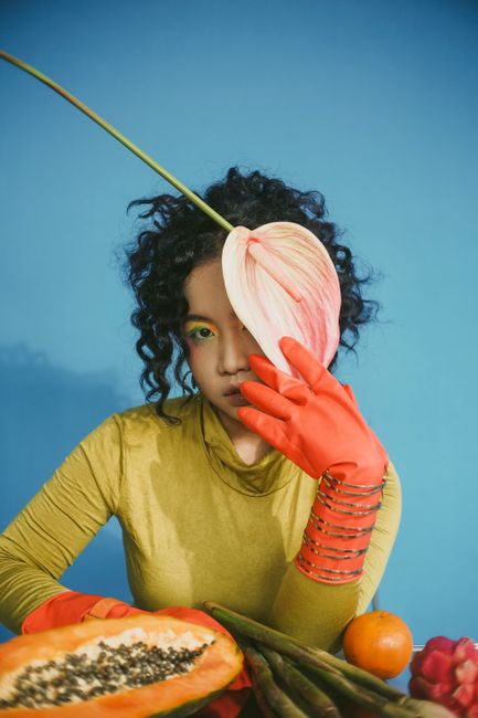 A woman posing with colorful fruits and plants against a vivid blue background.