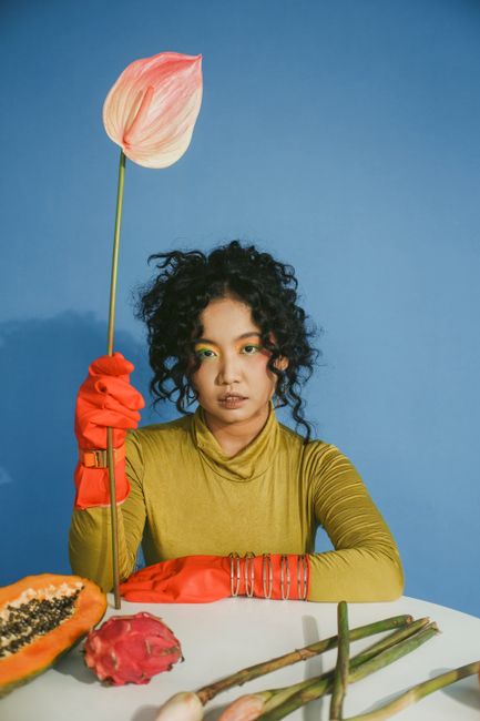 Bold portrait of a woman with a flower, orange gloves, and tropical fruits against a blue background.