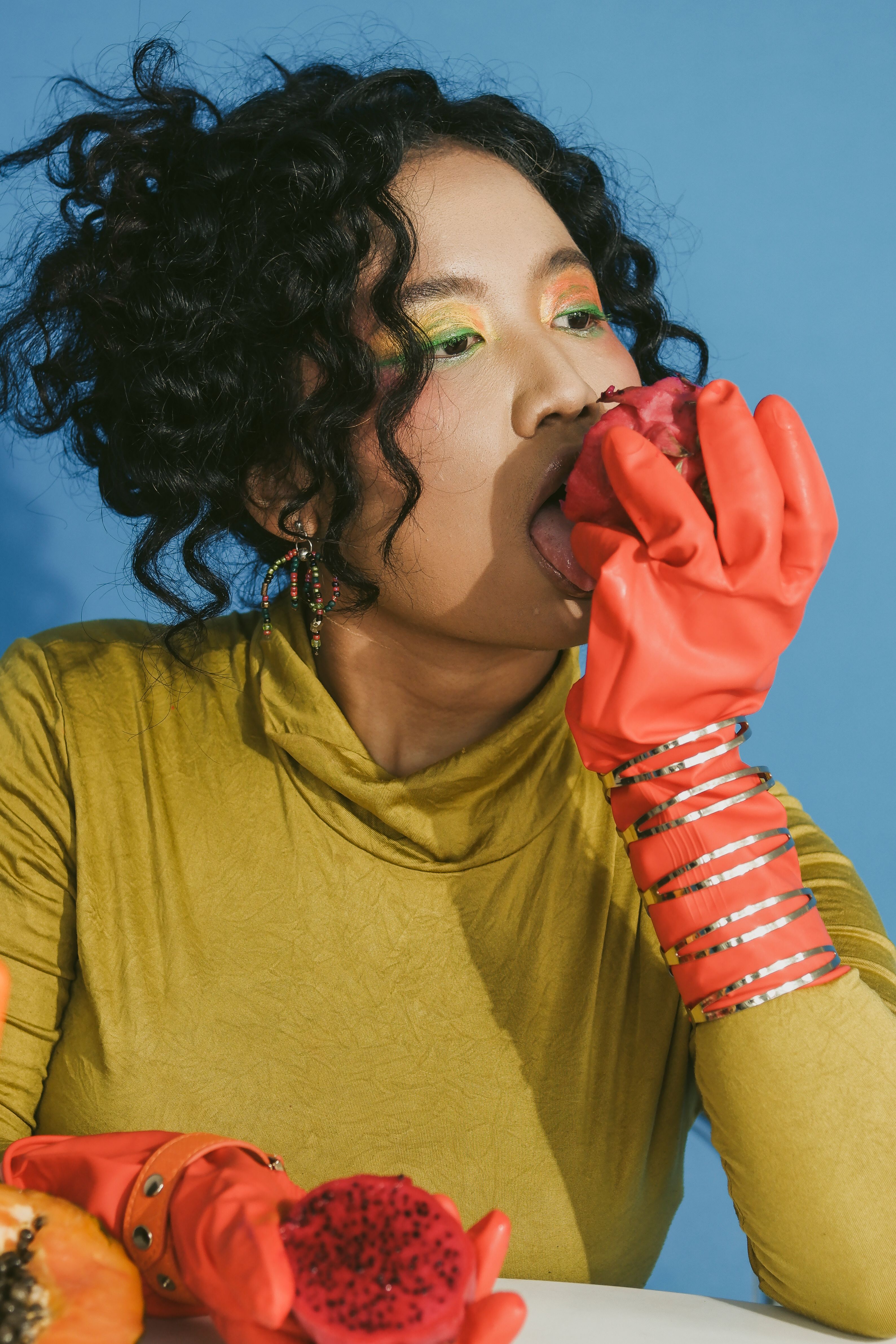 A fashionable woman in vibrant clothing enjoying a dragon fruit against a blue background.