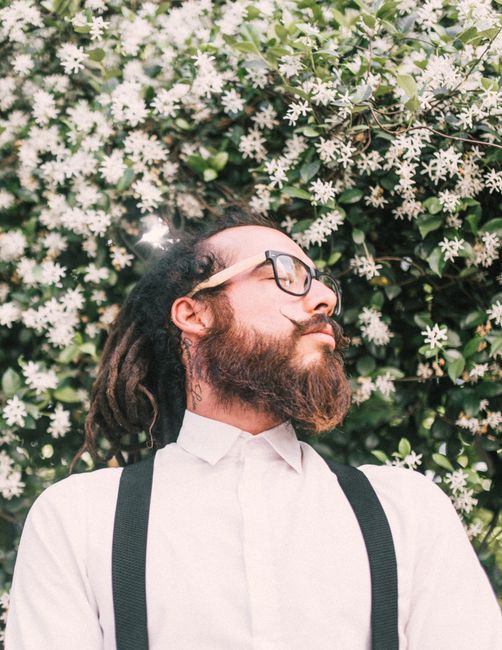 Stylish portrait of a bearded man with dreadlocks and glasses in front of white blossoms.