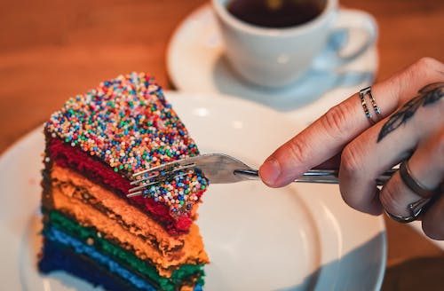 A person is holding a fork over a slice of rainbow cake