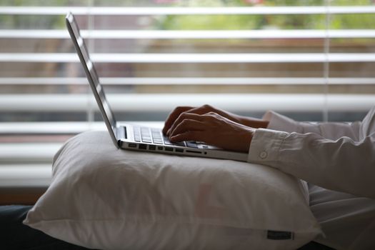Close-up of hands typing on a laptop while seated on a pillow indoors.