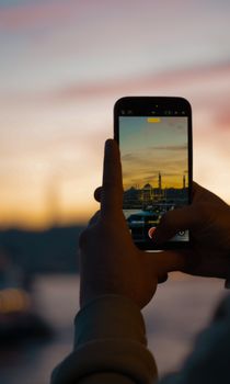Hands hold smartphone capturing Istanbul's skyline at sunset, featuring iconic landmarks and warm hues.