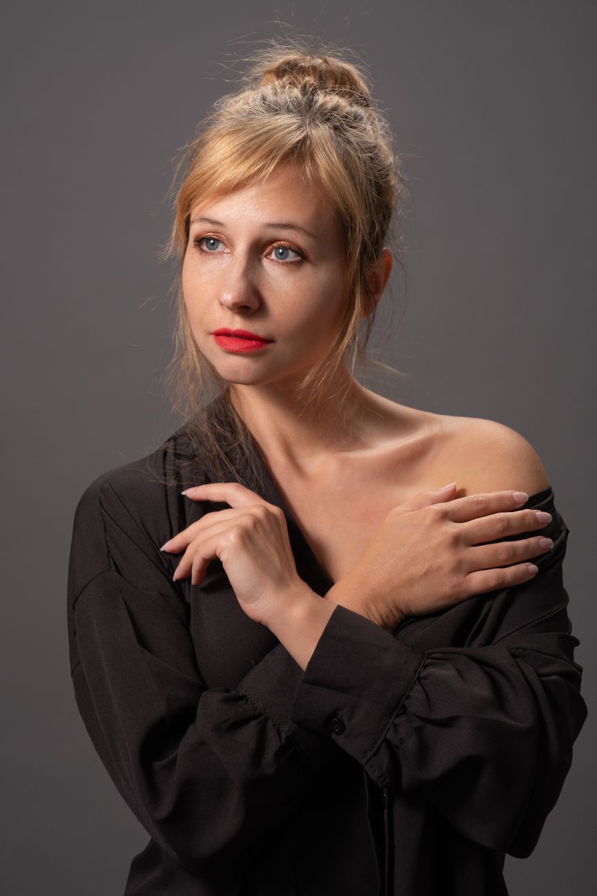 A blonde woman with red lipstick, crossing her arms in a studio portrait against a gray background.