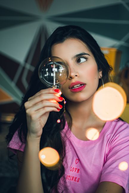 A young woman with dark hair and pink shirt looks through a light bulb indoors.