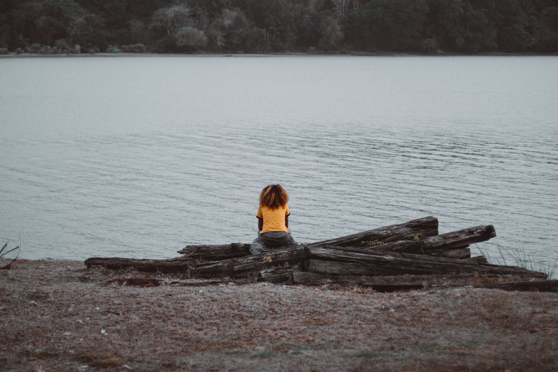 A woman in a yellow shirt sits alone on wooden logs by a calm lake, surrounded by nature.