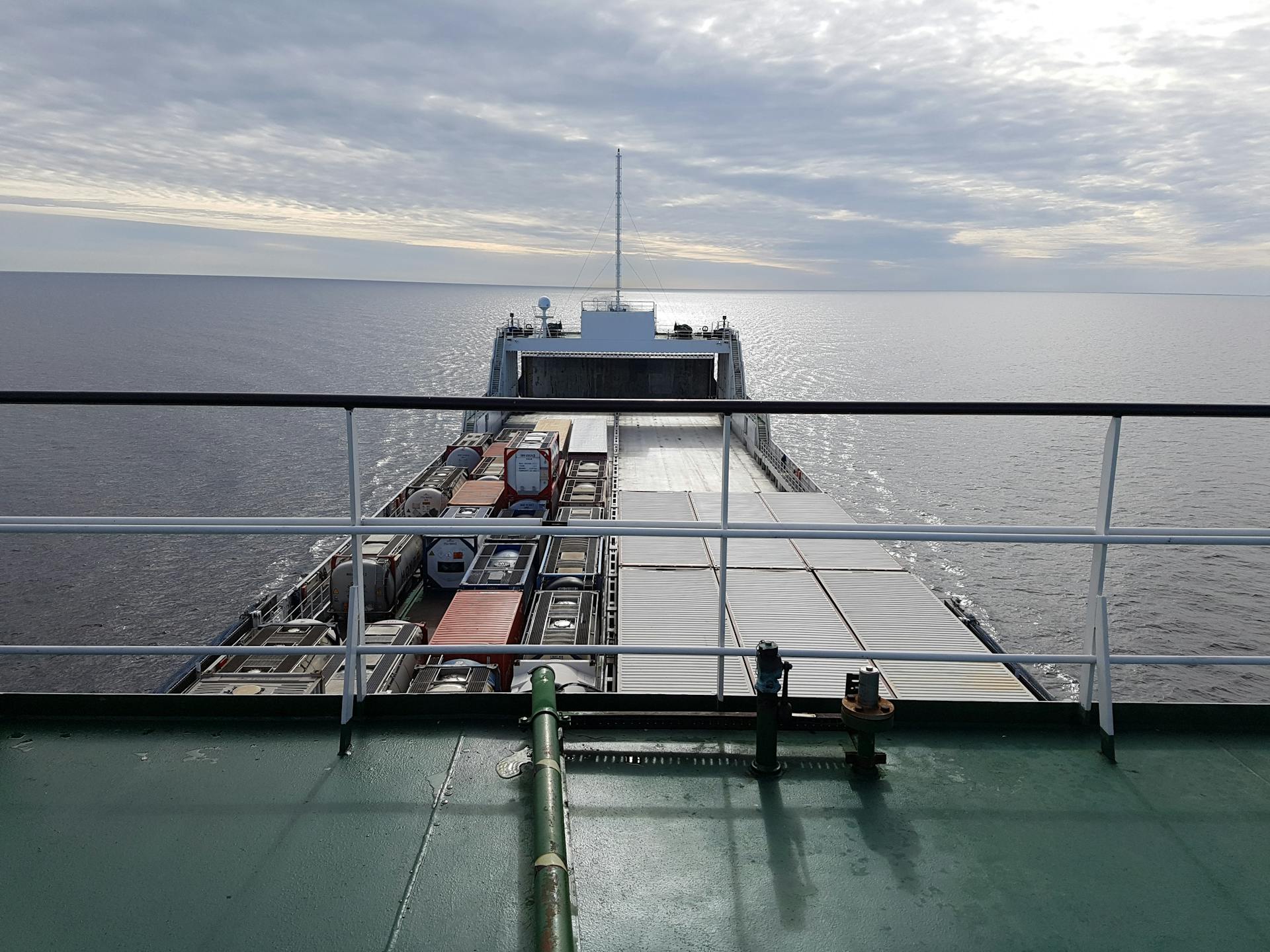 A large container ship sailing on the ocean under a cloudy sky, viewed from the deck.