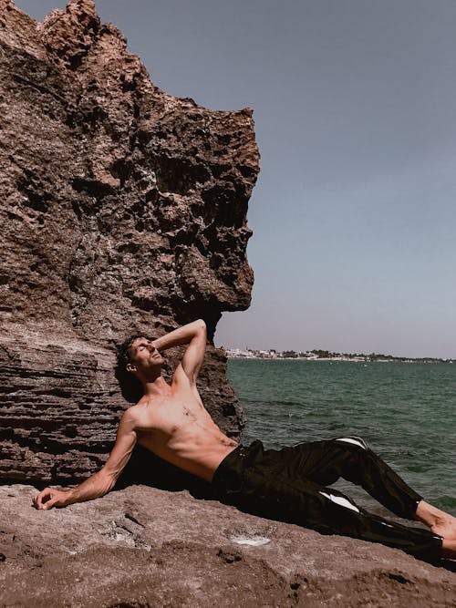 A shirtless man laying on a rock next to a body of water