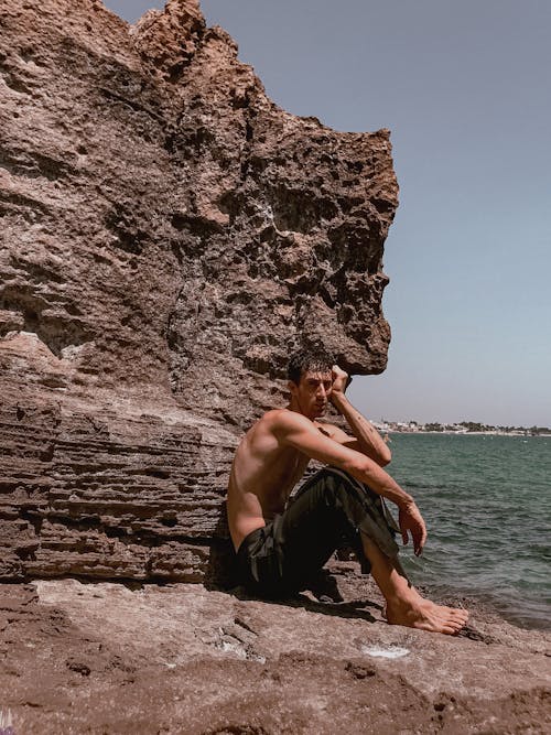 A shirtless man sitting on a rock near the ocean
