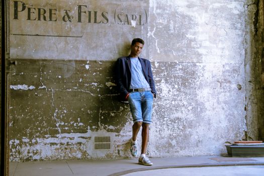 Stylish black man in denim shorts and suit jacket posing against a rustic wall.