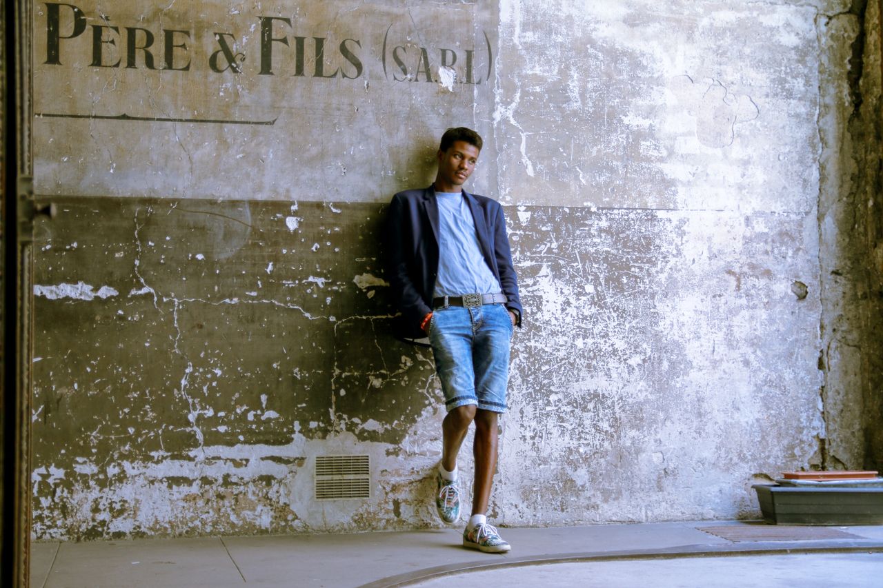 stylish black man in denim shorts and suit jacket posing against a rus
