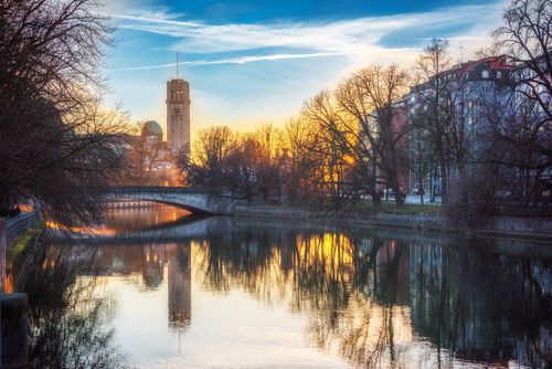 Free Scenic sunset view of Munich's riverside with a bridge, buildings, and trees reflecting in the water. Stock Photo