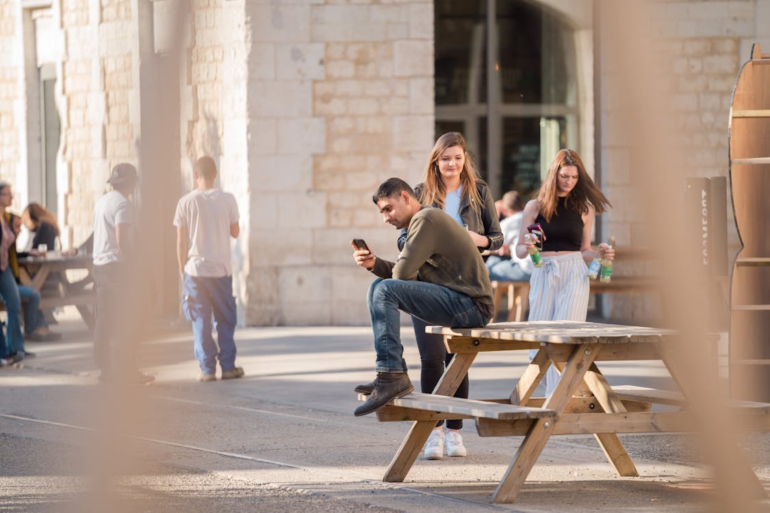 Free Man in Grey Long-sleeved Shirt and Blue Denim Jeans Sits on Picnic Table Near Women Walking Stock Photo