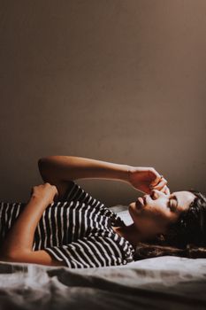 A woman lies relaxed in a sunlit bedroom, wearing a striped shirt.