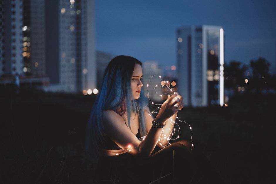 A woman sitting outdoors holding string lights against a cityscape backdrop during twilight.