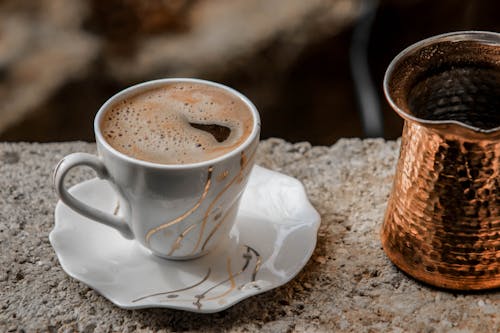 Close-up Photo of White Ceramic Cup and Saucer Set With Coffee Inside Next to a Brass-colored Coffee Pot