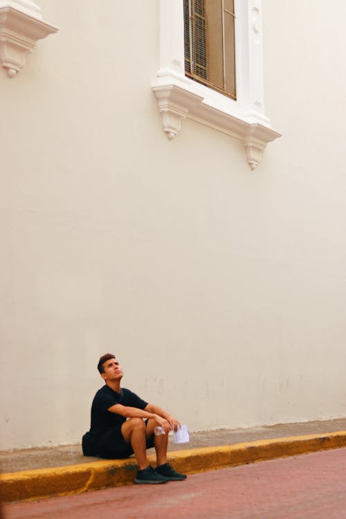 Free Man Sitting On Pavement Beside White Painted Wall Stock Photo
