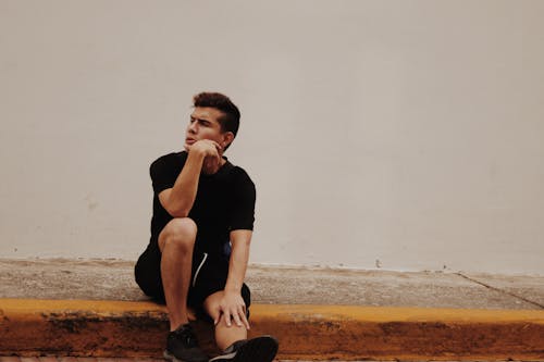 Free Photo of Man With Hand on Chin Sitting on Concrete Pavement In Front of White Wall Stock Photo